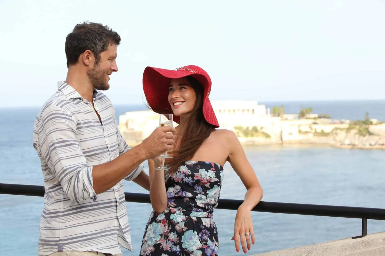 A man and woman stand on a balcony by the ocean. The woman wears a red hat and floral dress, holding a drink. The man is in a striped shirt, smiling at her.
