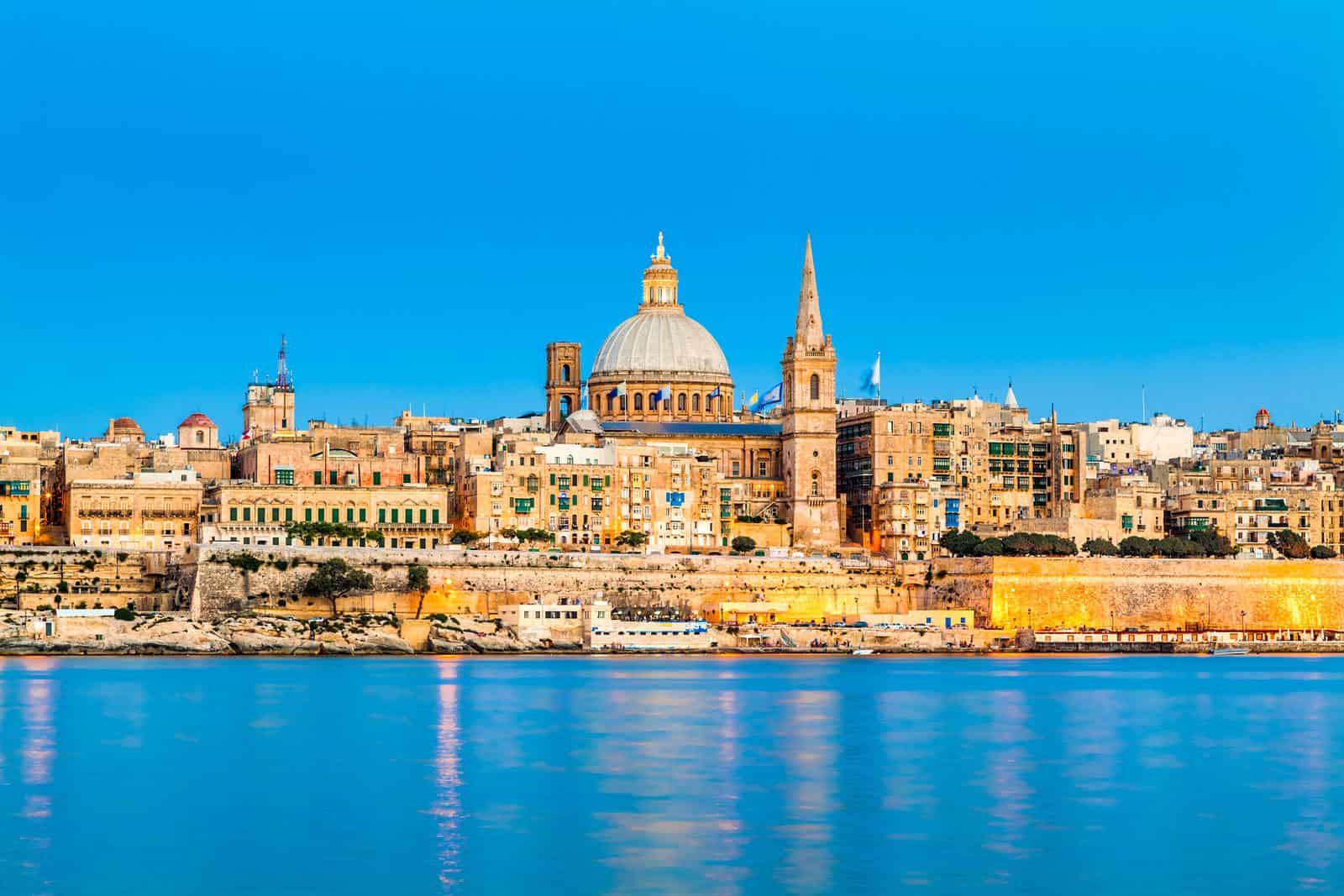 Skyline of Valletta, Malta, features stone buildings, a prominent domed church, and a tall spire, reflected in calm blue water under a clear blue sky.