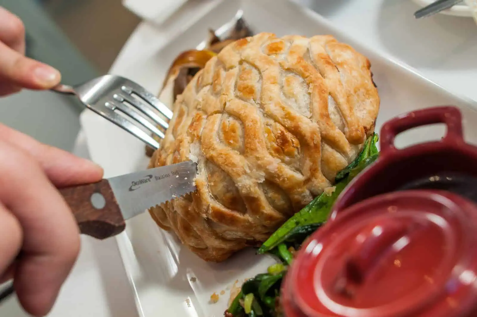 A person cuts into a pastry-covered dish on a white plate with greens on the side, using a knife and fork.