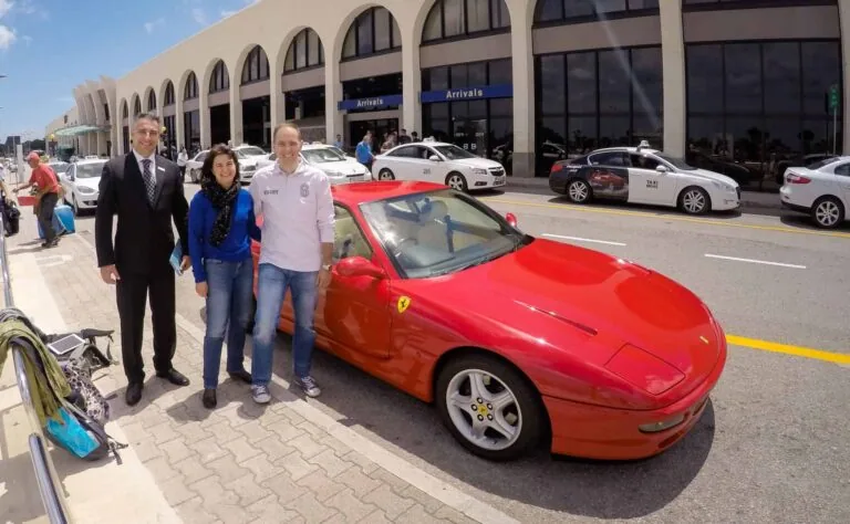Three people stand beside a red Ferrari parked in front of an airport terminal with taxis and an "Arrivals" sign in the background.