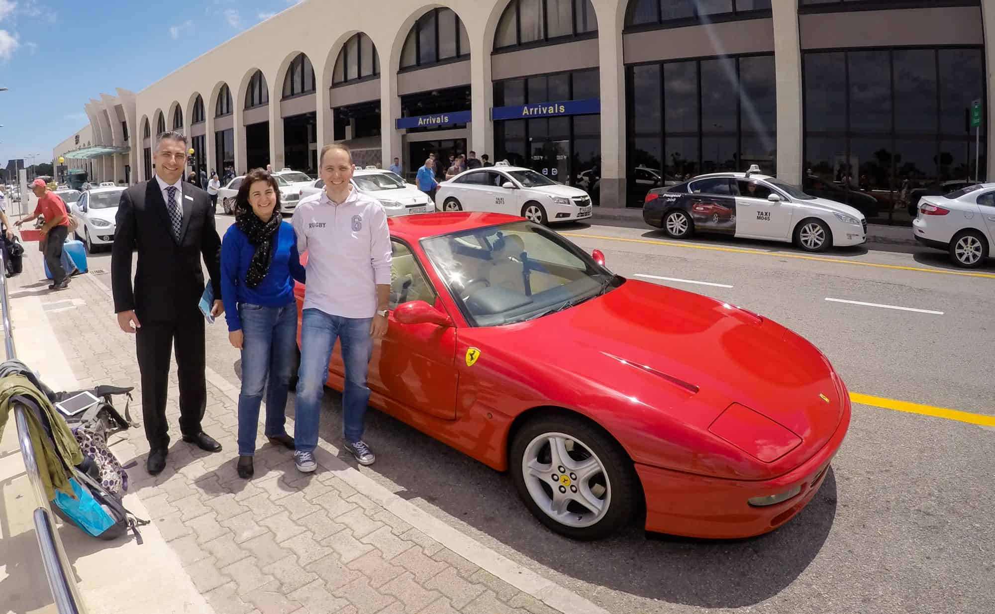 Three people stand beside a red Ferrari parked in front of an airport terminal with taxis and an "Arrivals" sign in the background.