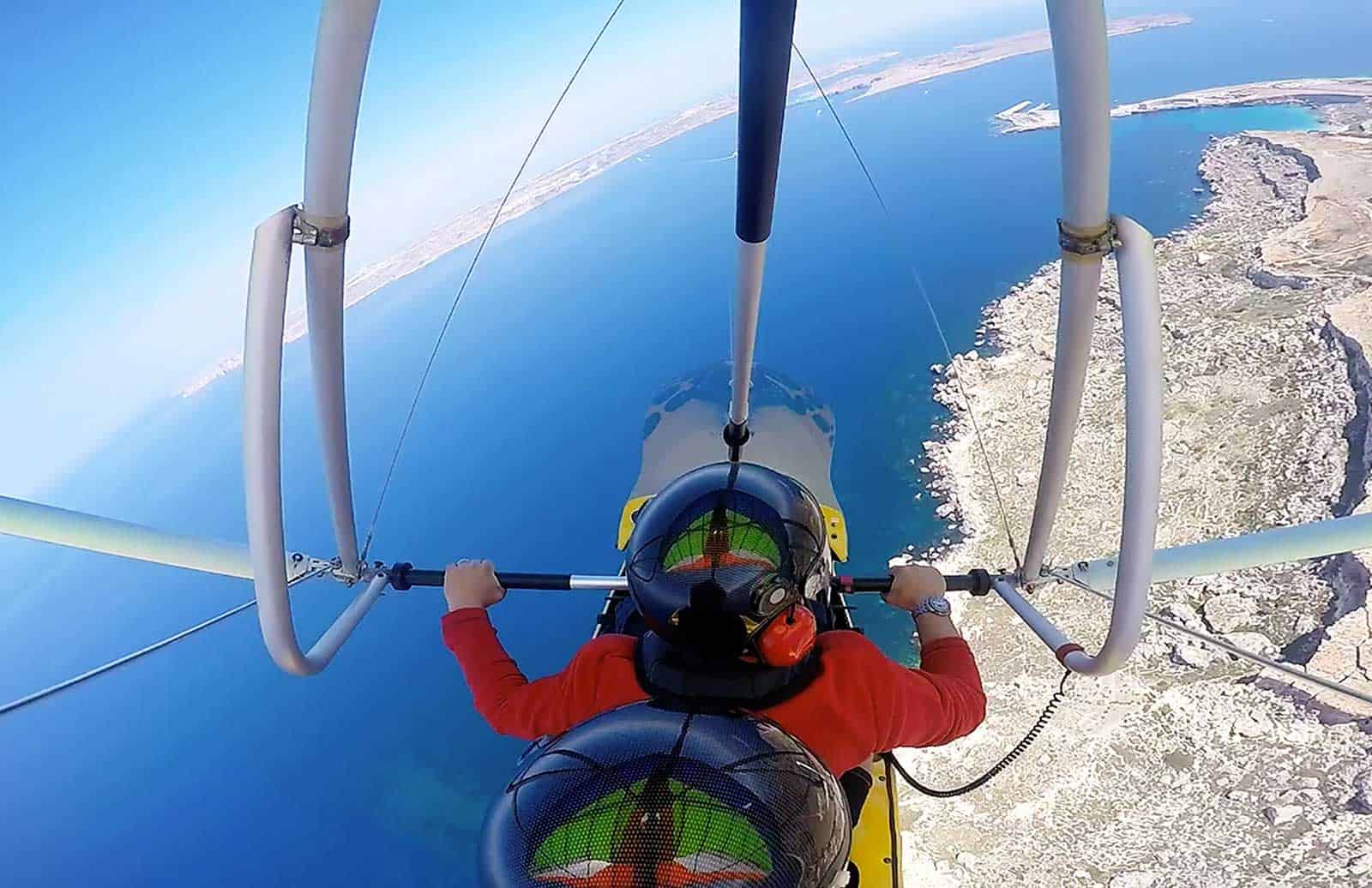 Two people in a hang glider flying over a coastline with blue water and rocky cliffs visible below.