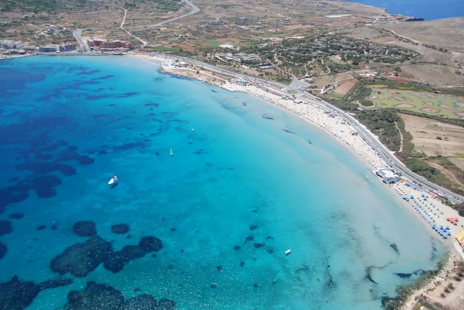 Aerial view of a coastal area with clear blue water, sandy beach, and scattered boats. Nearby are buildings and fields stretching into the background.