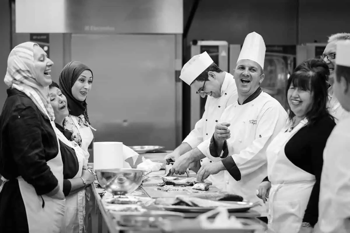 A group of chefs and participants, wearing aprons and chef hats, laugh and engage in a cooking class in a professional kitchen.