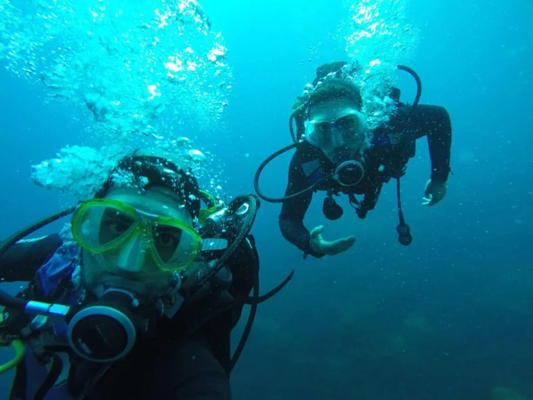 Two scuba divers underwater, surrounded by bubbles, wearing masks and breathing apparatus, with one person gesturing towards the camera.
