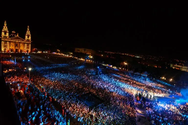 A large crowd gathers at night for an outdoor concert. The area is lit with colored lights, and a brightly lit building is visible on the left.