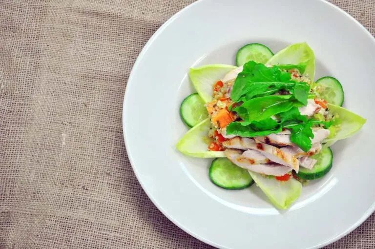 A white plate with a salad featuring sliced chicken, arugula, endive, diced vegetables, and cucumber slices, placed on a textured burlap surface.