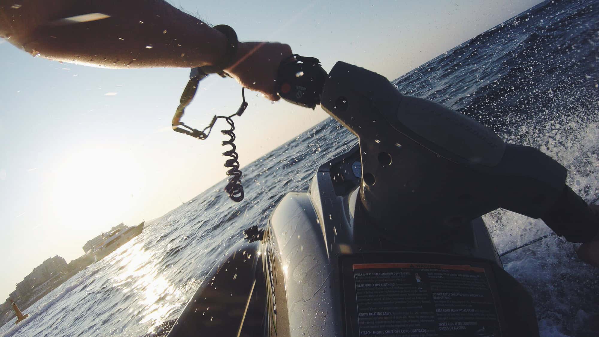 Person riding a jet ski on the ocean at sunset, holding the handlebars with a safety cord attached to their wrist.
