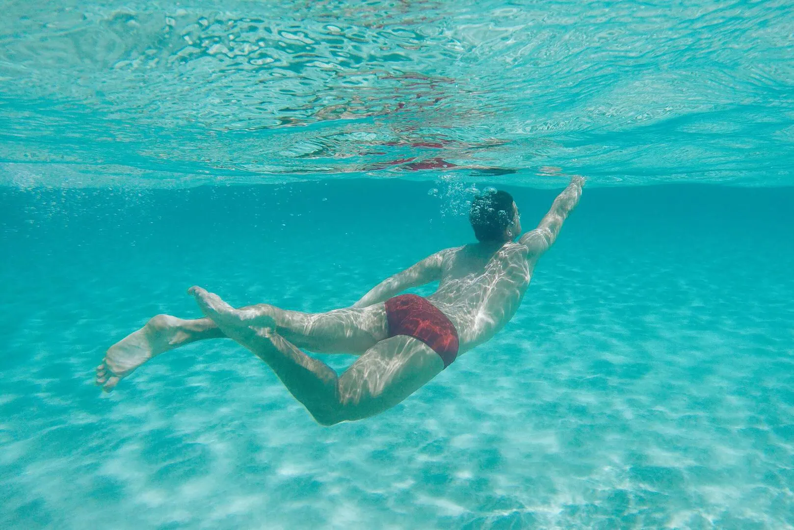Person swimming underwater in clear turquoise ocean wearing red swim trunks.