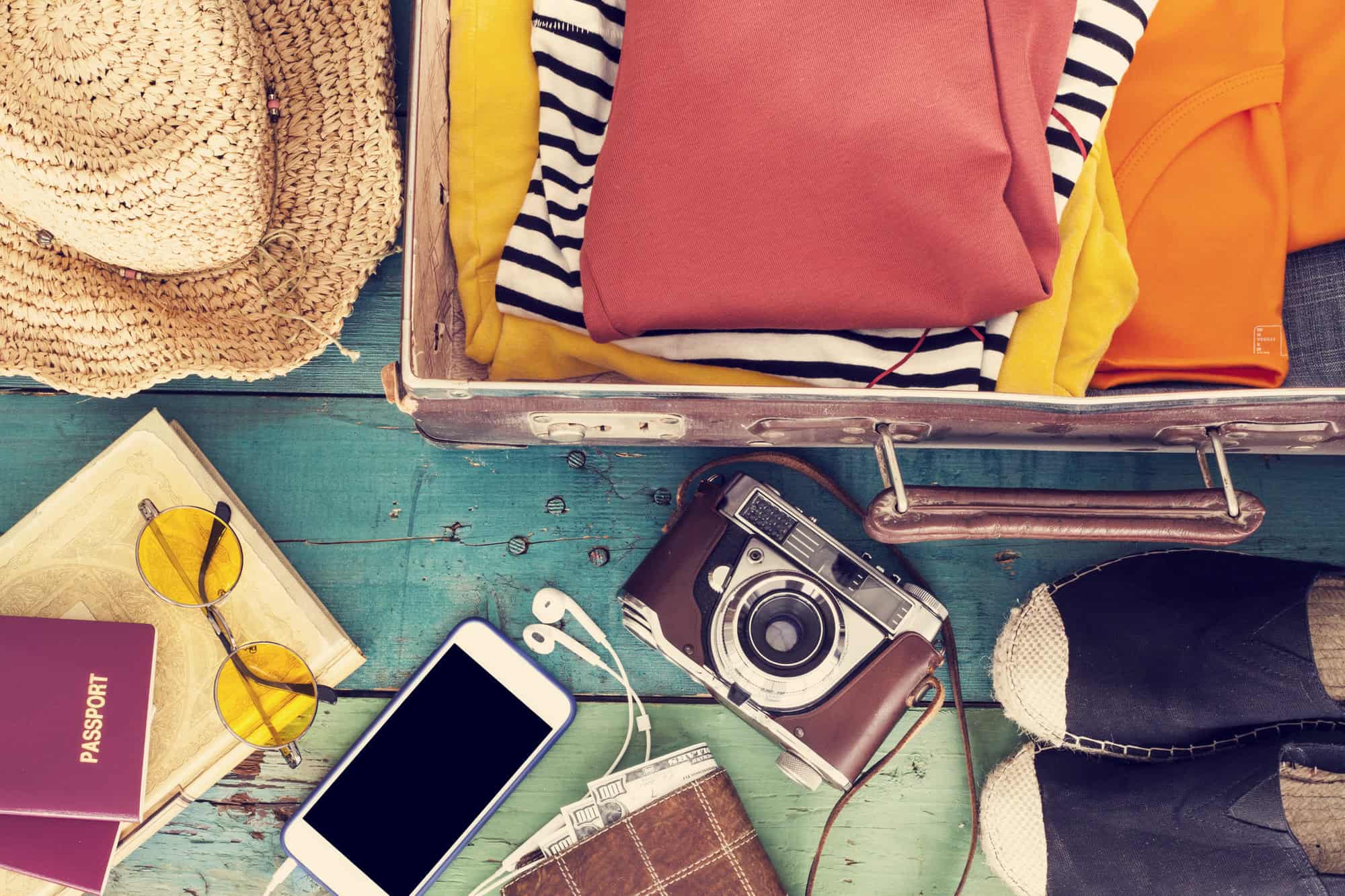 An open suitcase with folded clothes, a camera, hat, sunglasses, phone, earphones, a passport, and shoes on a wooden surface.