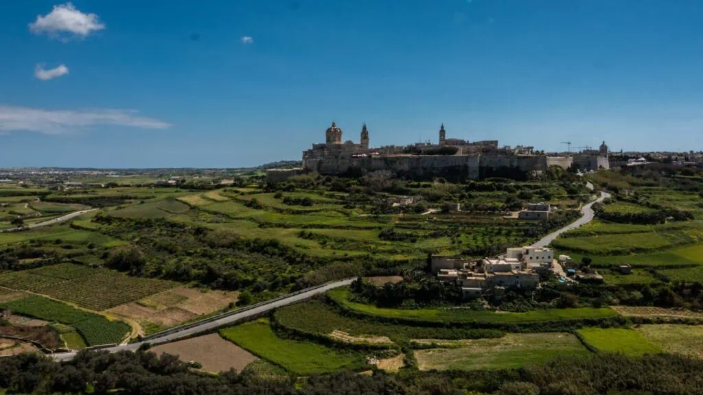 A scenic view of a fortified city on a hill surrounded by green fields and roads under a clear blue sky.