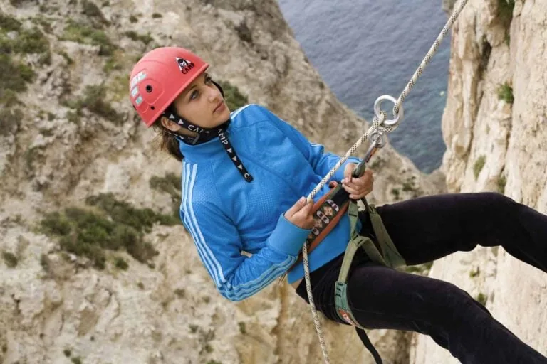 A person rock climbing with safety gear, wearing a red helmet and blue jacket, against a cliffside with water visible below.