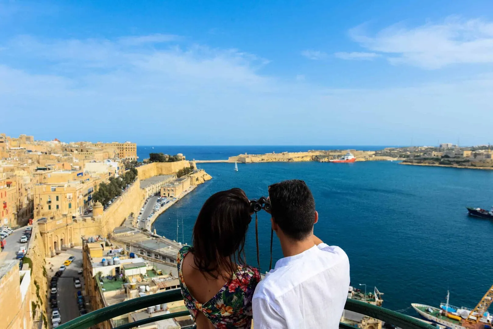 A couple stands on a balcony overlooking a coastal city with a calm, blue sea and boats. Buildings and roads are visible under a clear sky.
