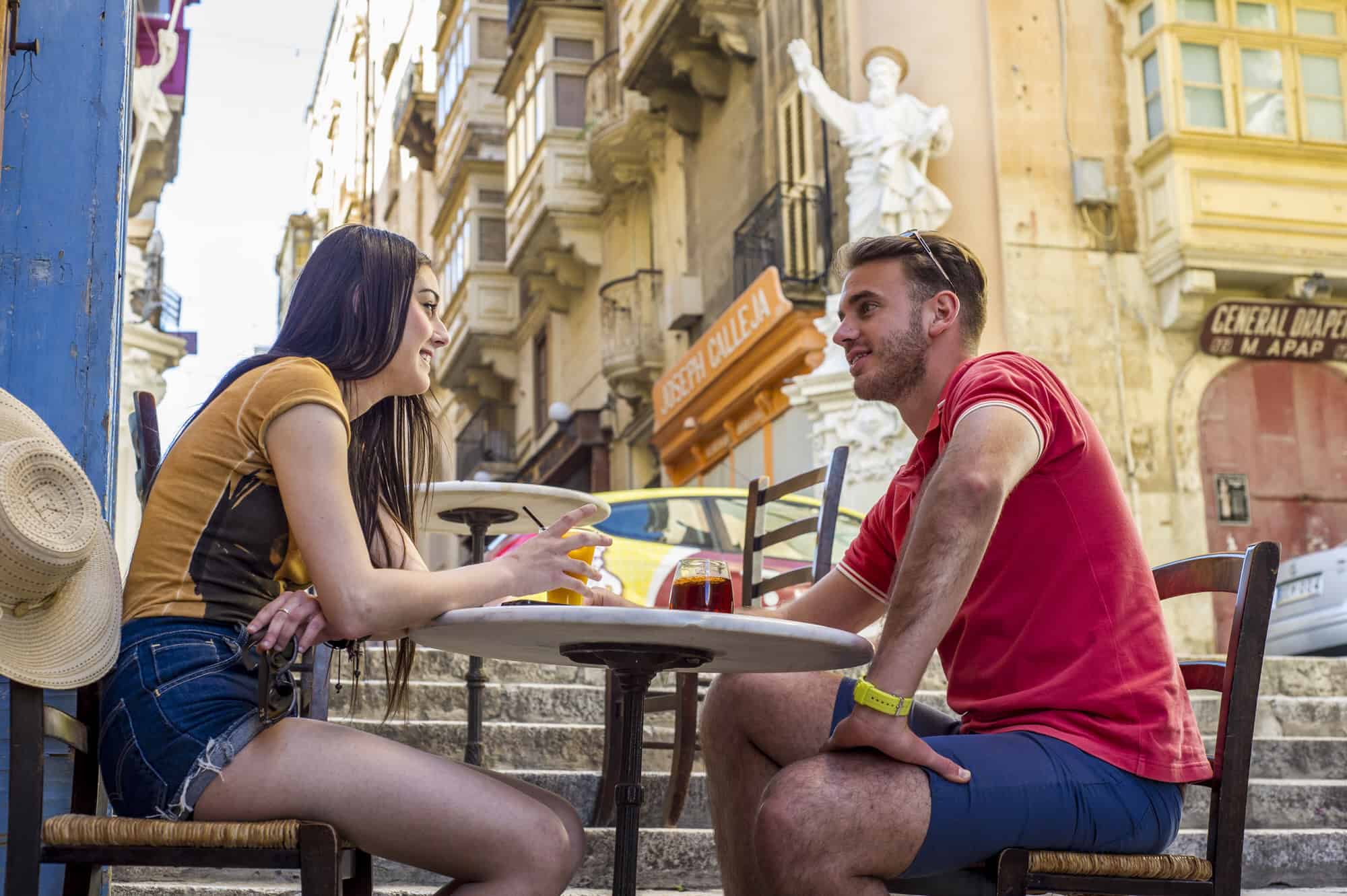 Two people sit at an outdoor cafe on a sunny day, engaged in conversation. A statue and old buildings are visible in the background.