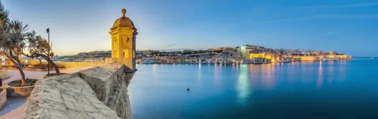 Panoramic view of a coastline with historic stone buildings overlooking calm blue water during sunset, featuring a watchtower on the left and a scenic bay extending to the horizon.