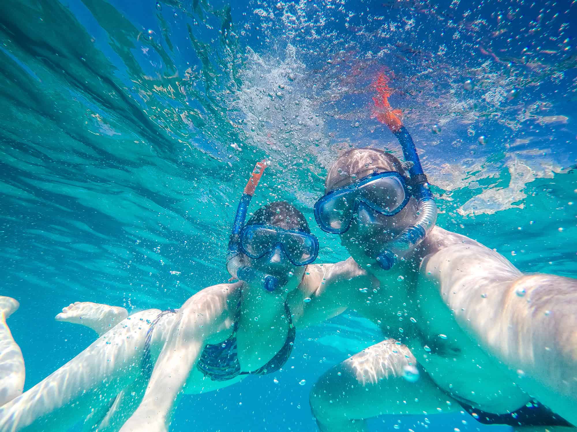 Two people wearing snorkeling gear underwater, surrounded by clear blue water and bubbles, taking a selfie while swimming.