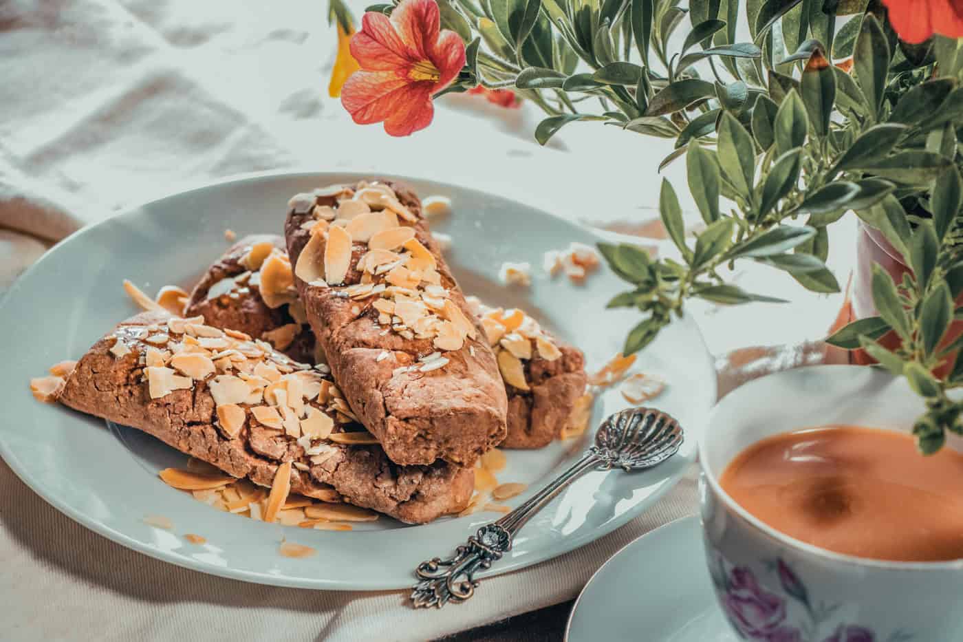 A plate with biscotti topped with almond slices next to a cup of coffee and a spoon, with a plant in the background.