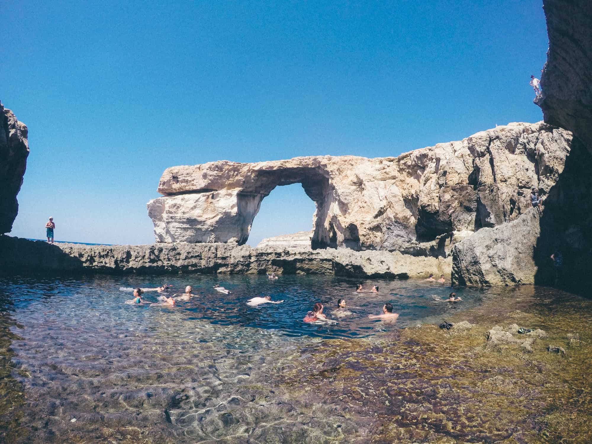 Swimmers in a natural pool near a rock arch formation under a clear blue sky.