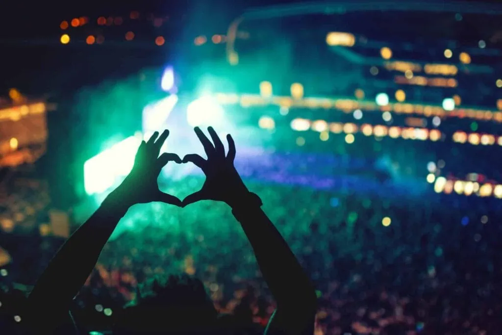 Silhouette of hands forming a heart shape against colorful lights and a large crowd at a concert.