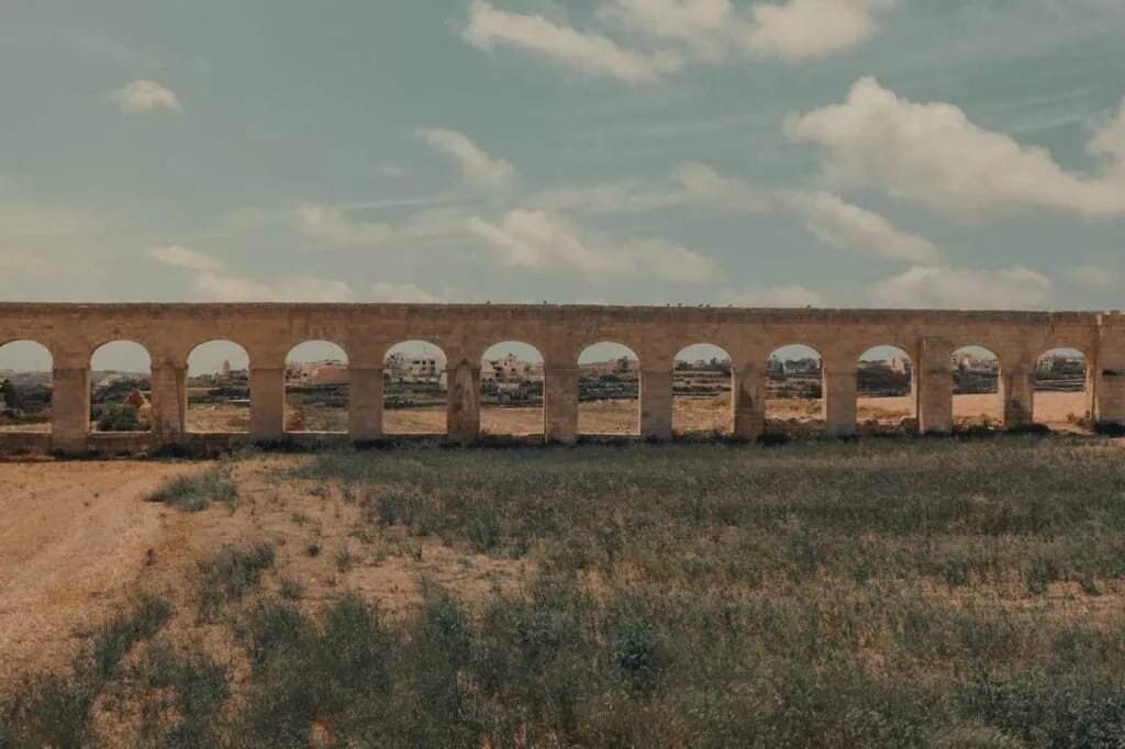 An ancient stone aqueduct with multiple arches stretches across a grassy field under a partly cloudy sky.