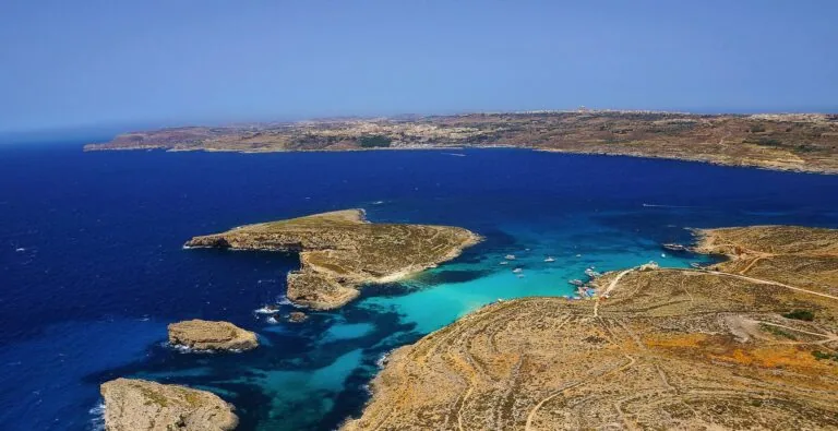 Aerial view of a coastline with brown rocky terrain and vibrant blue waters, featuring several small boats near the shore under a clear blue sky.