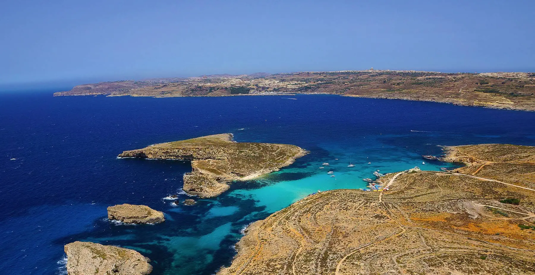 Aerial view of a coastline with brown rocky terrain and vibrant blue waters, featuring several small boats near the shore under a clear blue sky.