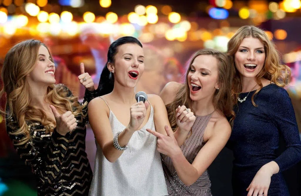 Four women smiling and pointing at a microphone, with a lively, blurred background of lights.