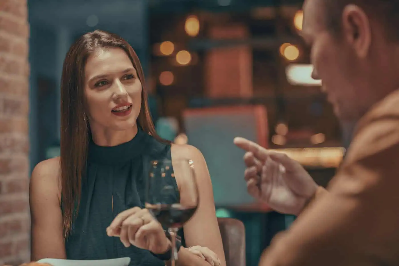 Two people sit at a table in a restaurant, engaged in conversation. A woman gestures while speaking; a wine glass is in front of her. The scene is warmly lit.