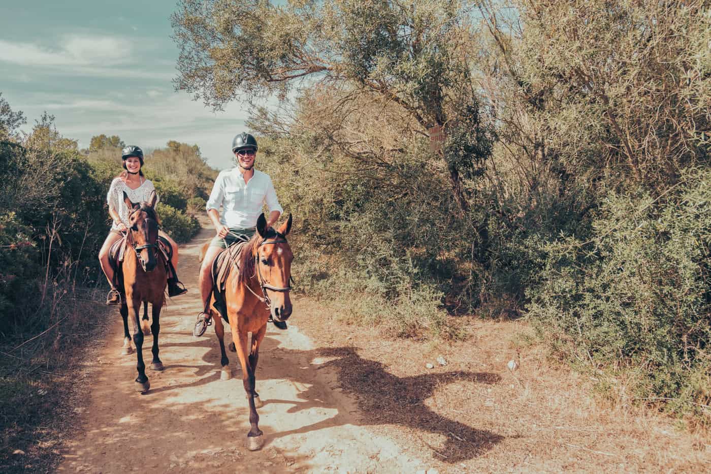 Two people riding horses on a dirt path through a wooded area, wearing helmets and casual attire.