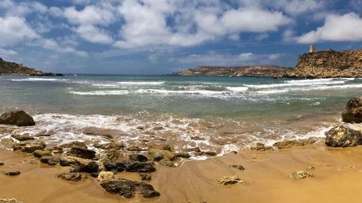 A sandy beach with scattered rocks, clear waves, and a cloudy blue sky. Cliffs and a tower are visible in the distance.