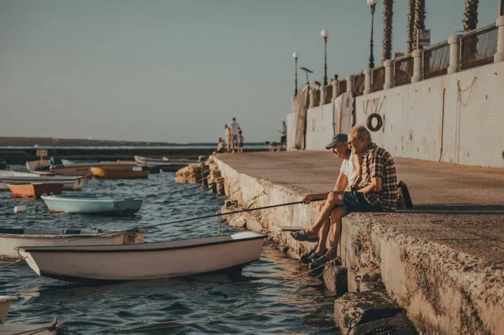 Two men sit on a stone pier fishing with rods in Malta. Several small boats float nearby in the calm water, while others stroll along the pier under a clear sky.