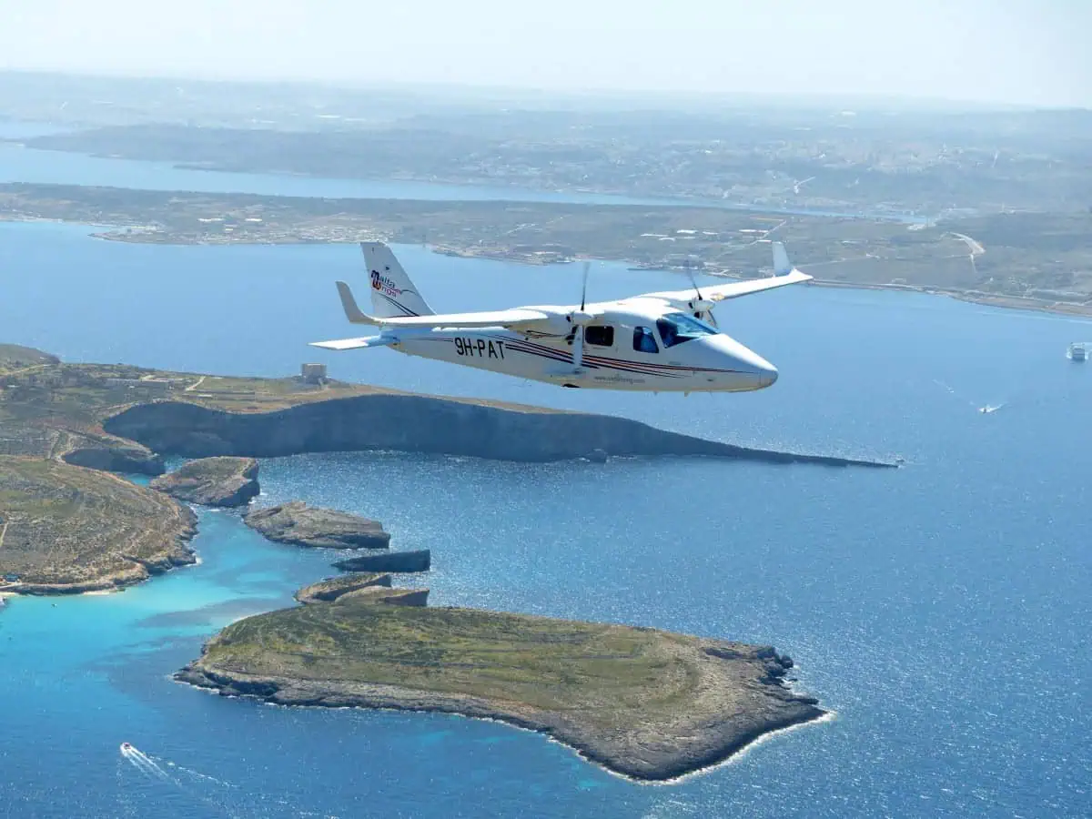 Small twin-engine plane flying over a vast blue sea with rugged coastlines and small islands below.