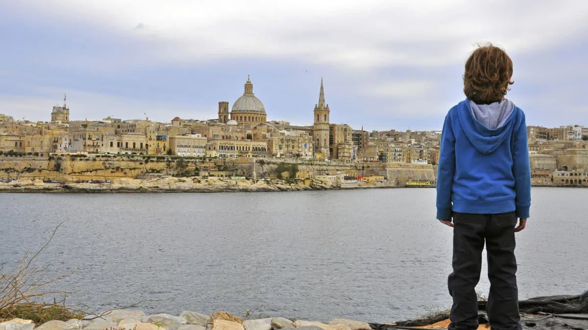 A child in a blue hoodie stands on a rocky shore, gazing across the water at Malta's skyline, where historic buildings and a prominent dome rise majestically. The cloudy sky above adds a touch of drama to this picturesque scene, perfect for a family adventure in Malta with the kids.