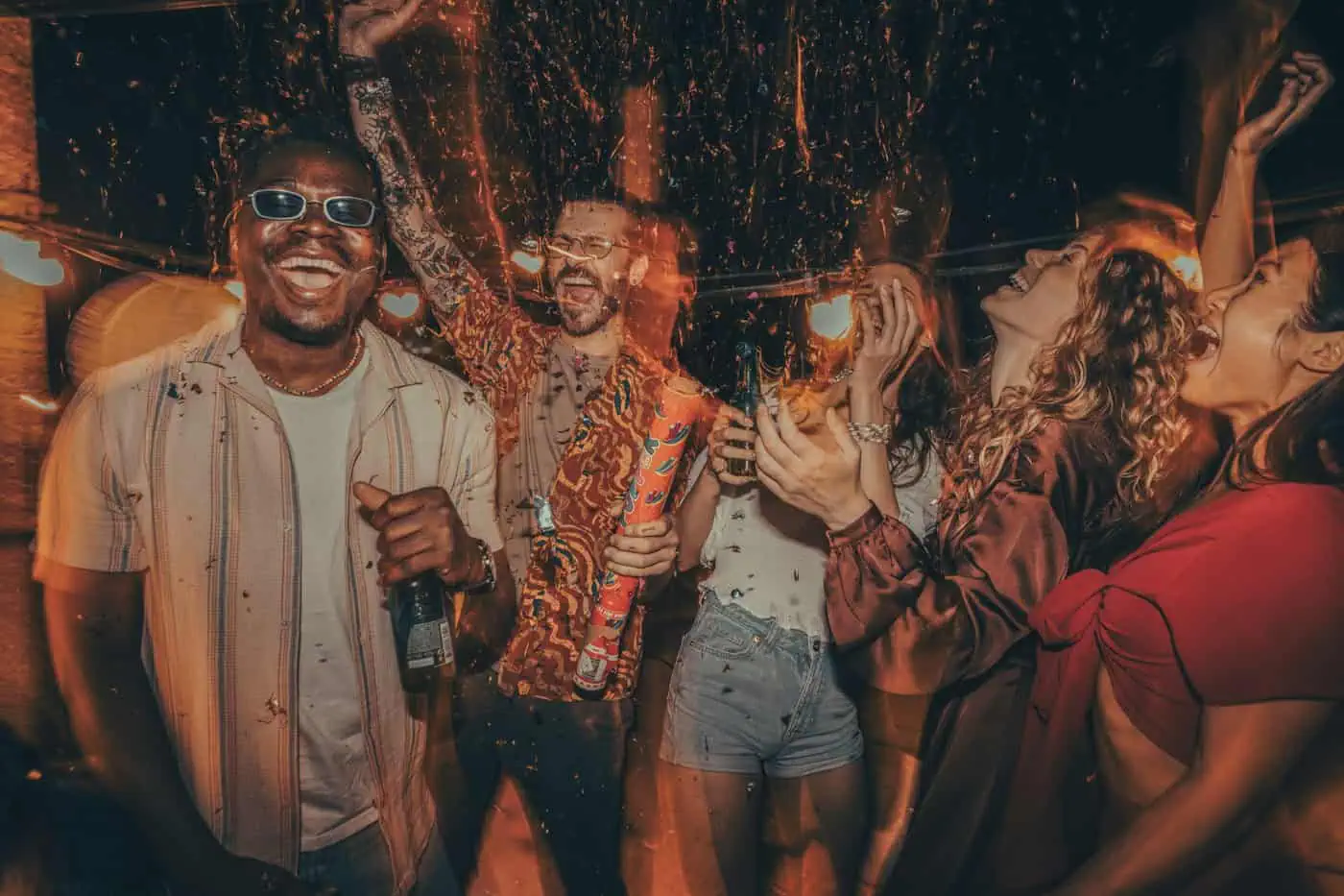 A group of people celebrating outdoors at night, holding drinks and smiling. String lights hang above, and one person is spraying confetti into the air.