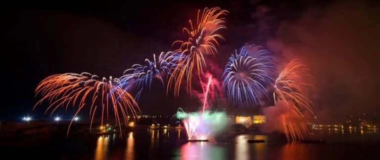 Colorful fireworks explode over a body of water, with reflections on the surface, against a dark night sky.