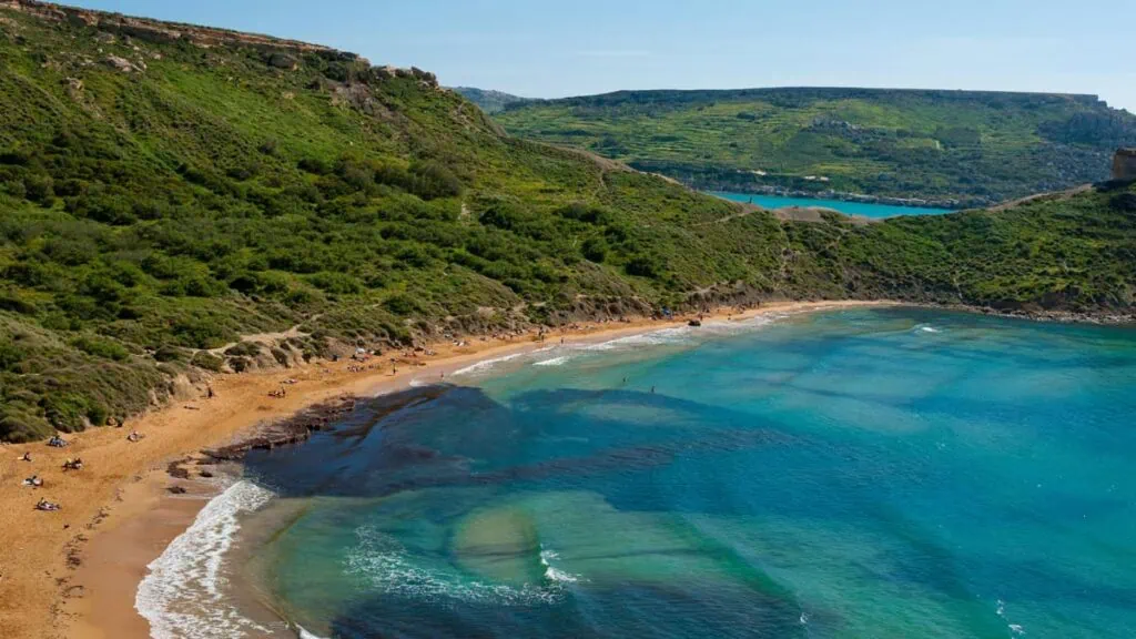 Aerial view of a beach with golden sand and clear turquoise water, surrounded by green hills under a clear blue sky. People are scattered along the shore.