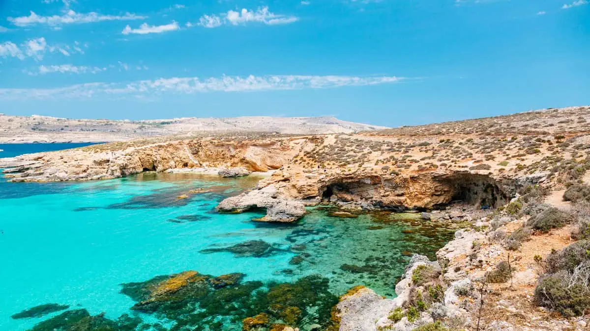 Coastal landscape with clear turquoise water, rocky cliffs, and sparse vegetation under a blue sky.