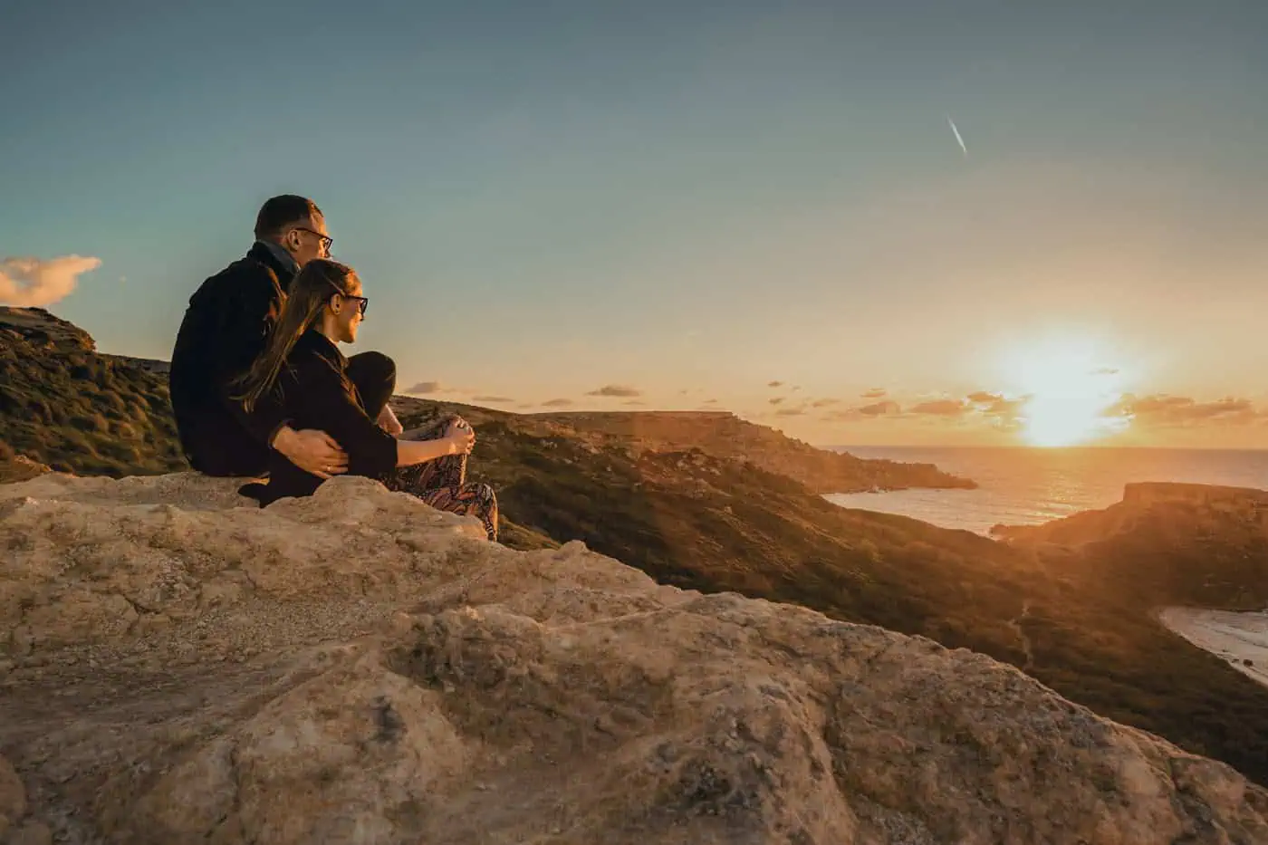 A couple sits on a rocky cliff, watching the sunset over the ocean with a clear sky.