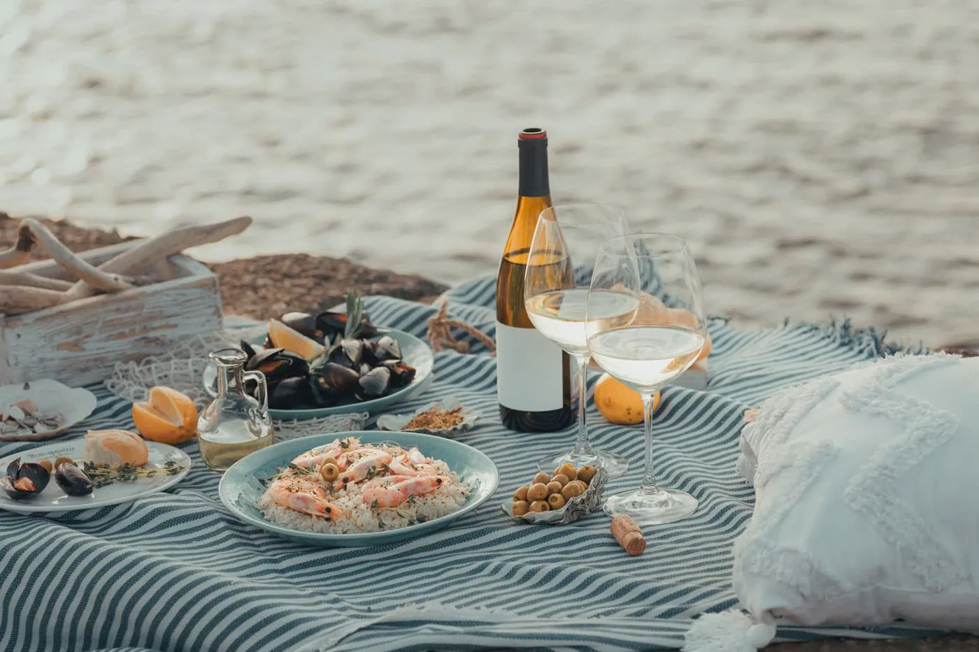 Picnic setup on a striped blanket with a seafood platter, mussels, wine bottle, two glasses of wine, olives, and lemons by the water.