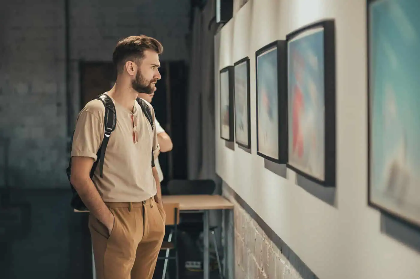 A man with a backpack observes framed artwork on a gallery wall.