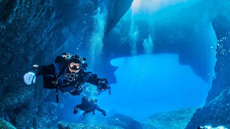Two divers explore an underwater cave surrounded by rocks, with clear blue water and sunlight filtering through.