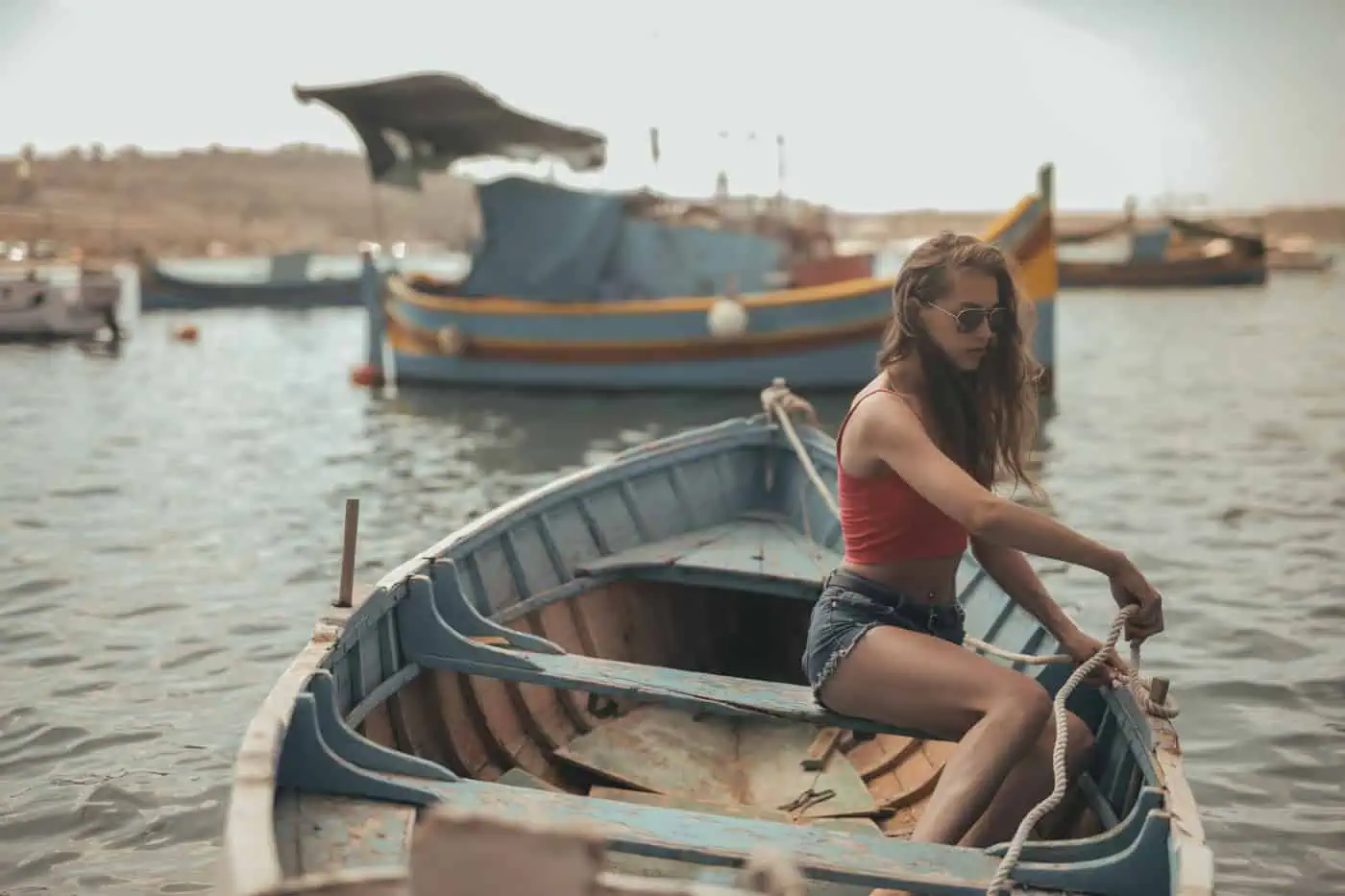 Woman in sunglasses sitting on a small blue wooden boat, holding a rope, with other boats in the background.