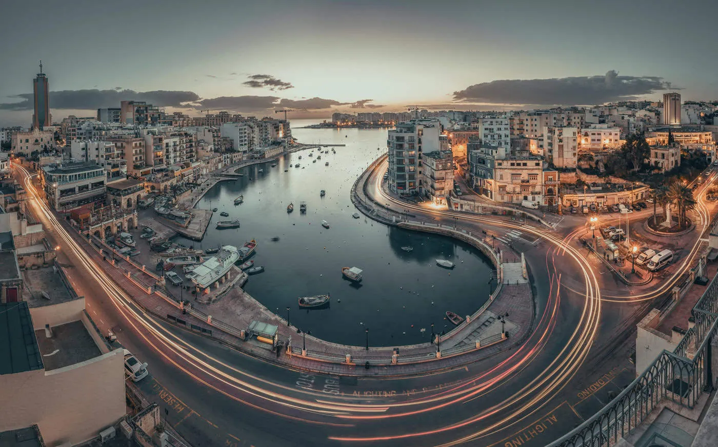 Panoramic view of a coastal city at dusk with cars leaving light trails around a curved waterfront. Buildings surround the harbor, and boats are moored in the water.