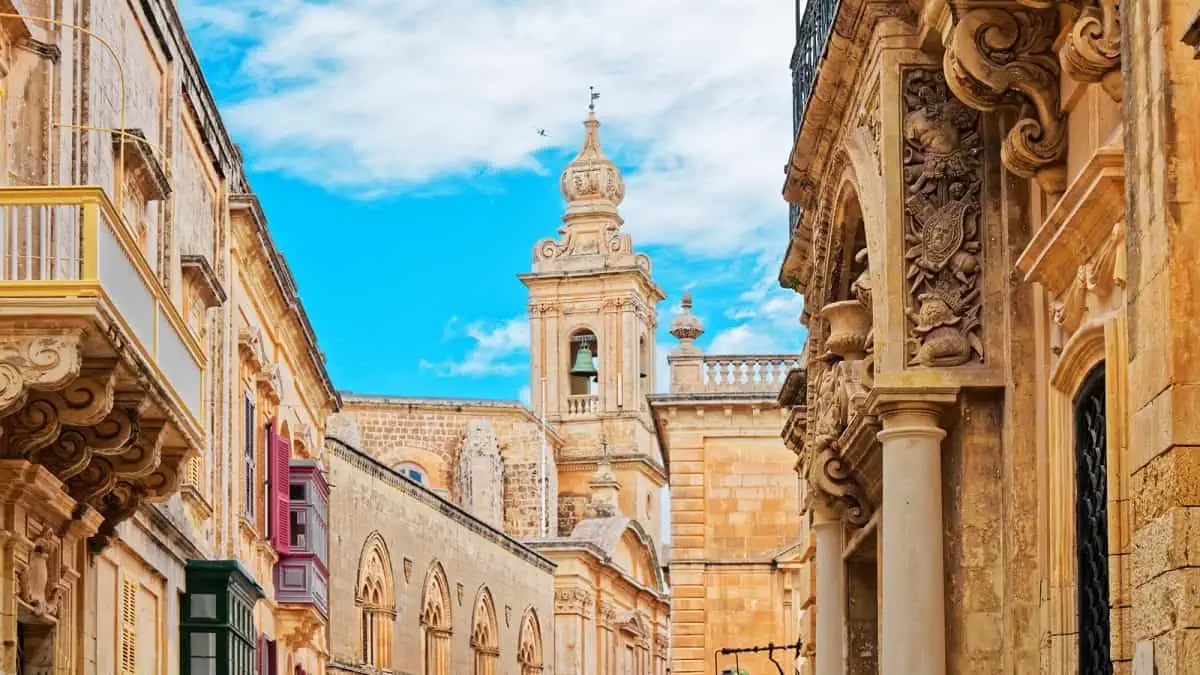 Historic stone buildings with ornate carvings and a central clock tower under a blue sky in a city scene.