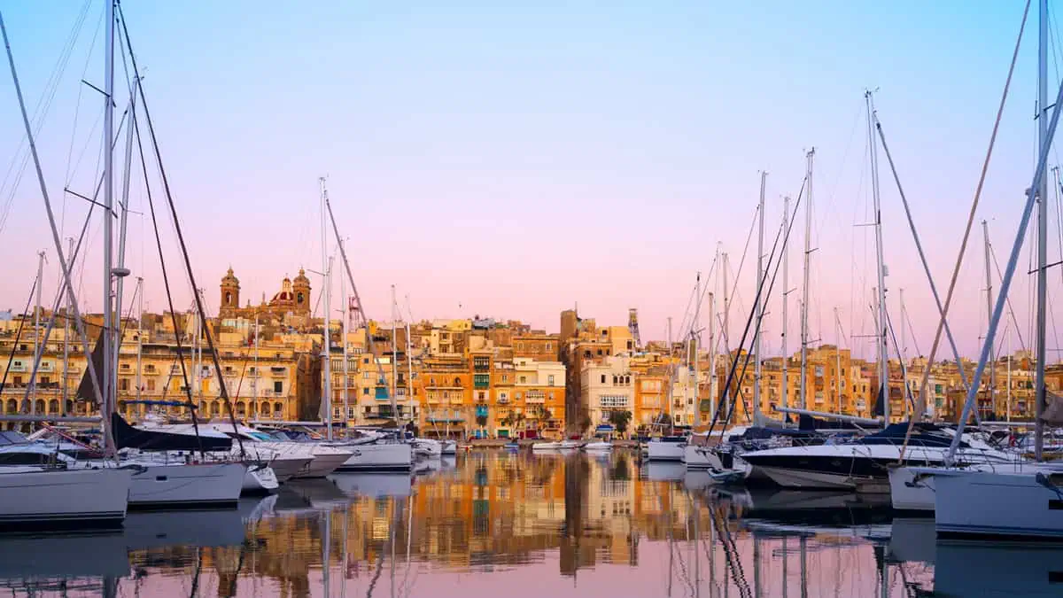 Harbor with sailboats docked in the foreground, reflecting on calm water at sunset. A historic town with warm, yellow-lit buildings is visible in the background under a pinkish sky.