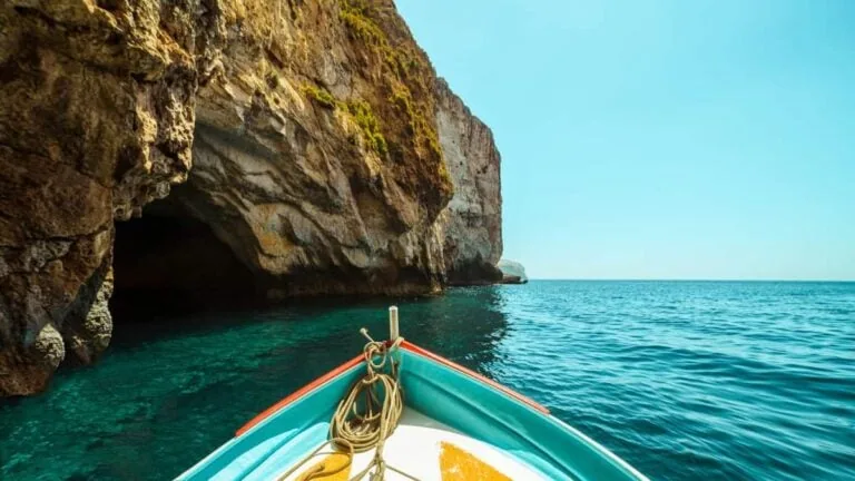 Boat approaching a rocky cave entrance on a clear blue sea under a bright sky.