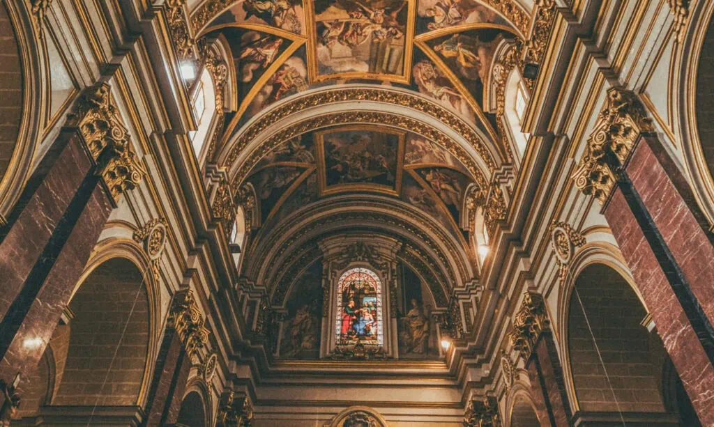 Atmospheric churches in Malta boast ornate interiors with arched ceilings, detailed murals, and stunning stained glass windows that depict captivating religious scenes, all viewed from ground level.