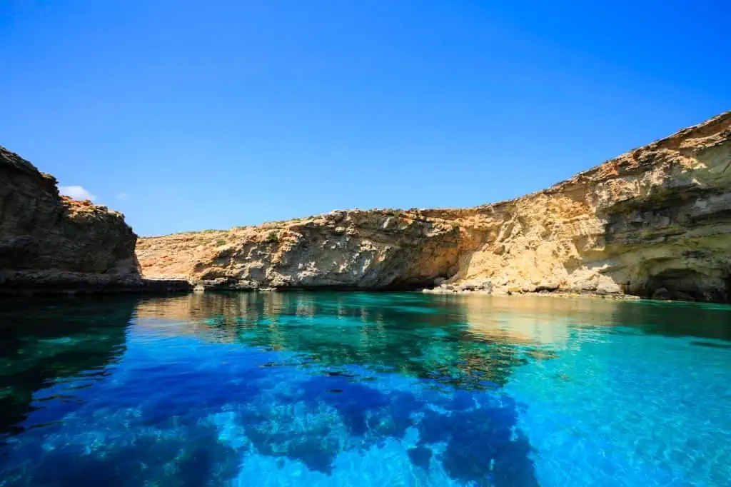 Clear blue water with rocky cliffs under a bright blue sky, reminiscent of the atmospheric charm found near churches in Malta.