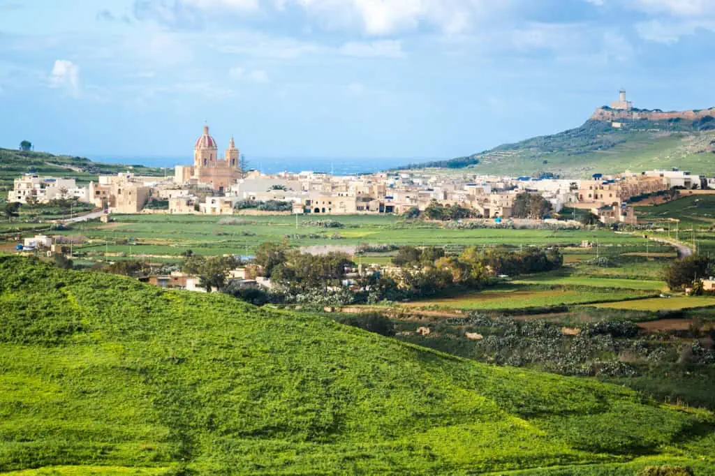 Nestled among lush green fields and rolling hills, an atmospheric Maltese village is marked by its prominent domed church under a cloudy sky.