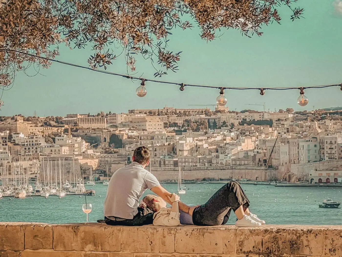 A couple relaxes on a stone ledge overlooking a waterfront cityscape, with decorative lights hanging above, evoking the charm of atmospheric churches in Malta.